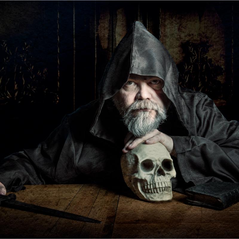 A monk leans on a human skull and holds a dagger.